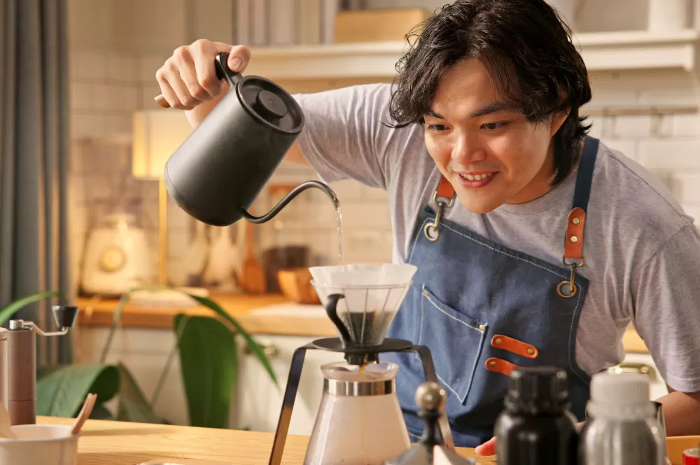 A man smiling as he pours water from a gooseneck kettle into a glass V60 dripper.