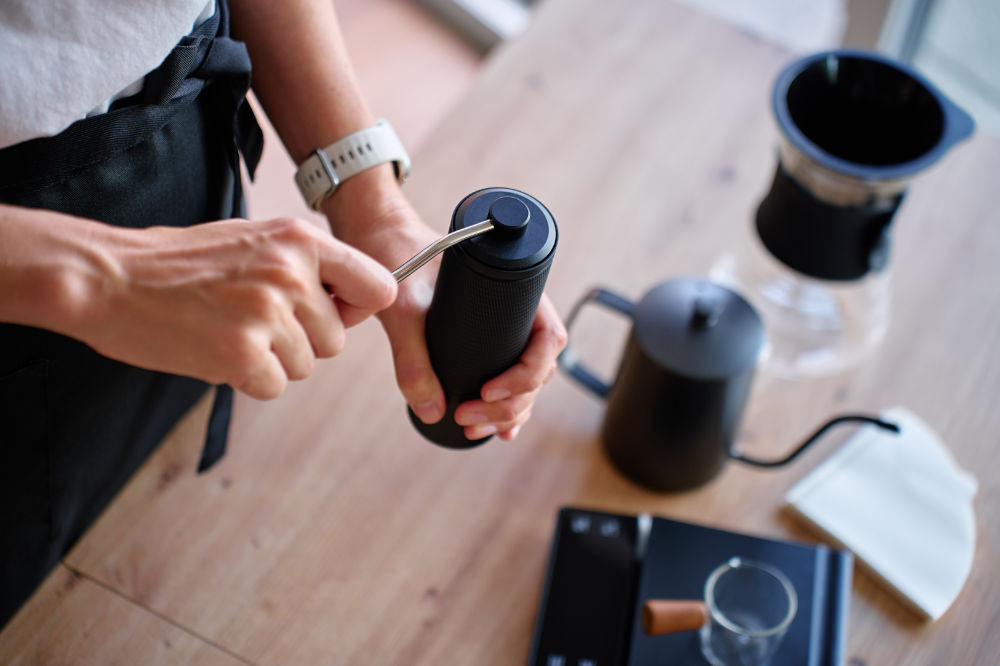 Hands turning a black hand grinder, with a kettle, dripper and scale set out on the table.