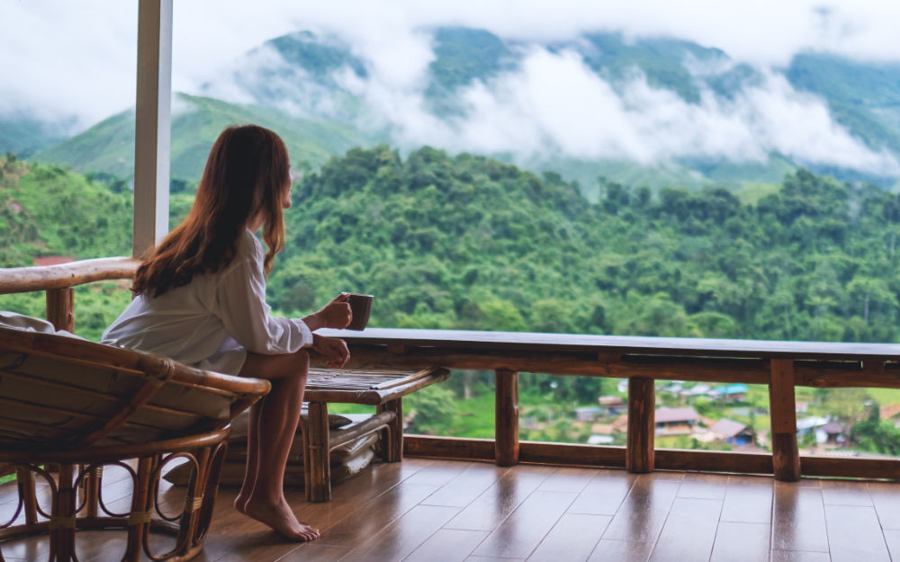 A woman with a cup of coffee on a wooden balcony, looking out over misty green mountains.